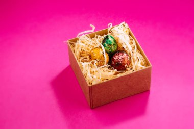 Kırmızı, yeşil ve sarıya boyanmış üç Paskalya yumurtası içinde ahşap talaşlar bulunan bir kutunun içinde yatıyor. Pembe arka planda boyanmış paskalya yumurtaları. Boşluğu kopyala