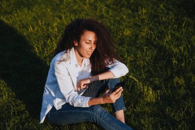 A young girl sits on the green grass in the park and uses social networks using a mobile phone.