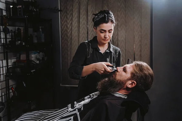 Young woman barber making haircut of bearded man in barbershop. Self-care, masculine beauty. Hair care service concept