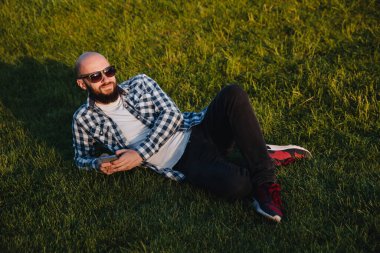 A young man sits on the green grass in the park and uses social networks using a mobile phone.