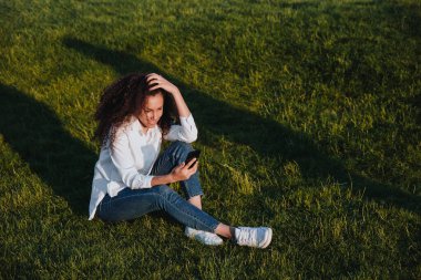 A young girl sits on the green grass in the park and uses social networks using a mobile phone.