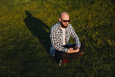 A young man sits on the green grass in the park and uses social networks using a mobile phone.