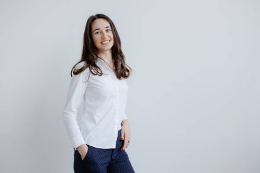 Genç bir kadın, bir eli cebinde açık gri bir duvara karşı dururken parlak bir şekilde gülümsüyor. Kendine güveni tam, beyaz gömlek ve koyu mavi pantolon giyiyor..