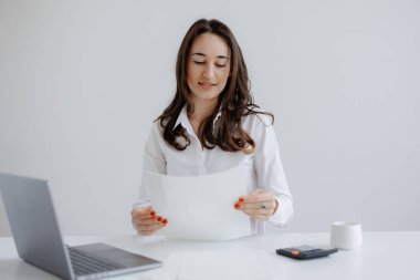 Bir kadın masasında evrakları incelerken gülümsüyor. Yanında dizüstü bilgisayar ve hesap makinesiyle aydınlık bir ofiste oturuyor. Odaklanmış ama rahat bir atmosfer yaratıyor..