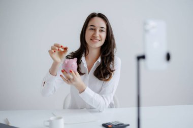 Kumbarayı tutarken uzun saçlı bir kadın gülümsüyor ve sanal bir buluşma ortamında kameraya gösteriyor. Atmosfer parlak ve profesyonel. Finansal tartışmalar için ideal..