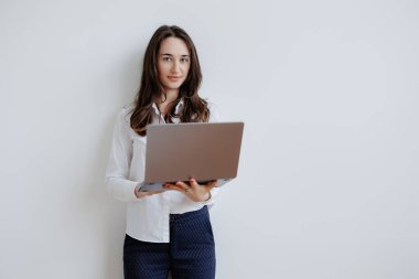 Temiz bir duvara dayanmış genç bir kadın, elinde bir dizüstü bilgisayarla doğrudan kameraya bakıyor. Meşgul ve fikirlerini paylaşmaya ya da profesyonel bir ortamda işbirliği yapmaya hazır görünüyor..