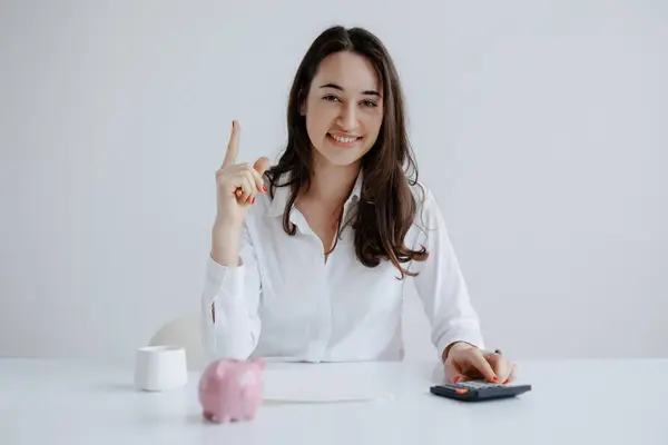 Elinde kumbarası ve hesap makinesiyle masada oturan bir kadın yukarı bakarken neşeli görünür. Parlak, minimalist atmosfer onun pozitif davranışını arttırır..