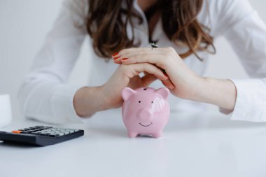 Beyaz gömlekli bir kadın, ellerini pembe bir kumbara üzerine koyarak finansal planlarına odaklanıyor. Yakınlarda temiz bir masanın üzerinde, bütçe faaliyetlerini gösteren bir hesap makinesi bulunmaktadır..