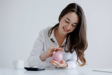 Uzun saçlı bir kadın, temiz bir masanın üzerindeki pembe kumbaraya bozuk para koyarken gülümsüyor. Güneş ışığı çalışma alanını aydınlatıyor, sıcak ve davetkar bir atmosfer yaratıyor..