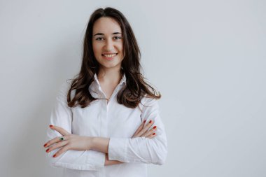 Genç bir kadın kollarını kavuşturmuş, sıcak bir şekilde gülümsüyor. Uzun saçları yavaşça omuzlarına dolanıyor ve beyaz bir gömlek giyiyor..