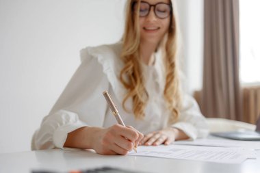 Beyaz bluz giymiş genç bir kadın, pencereden gelen yumuşak, doğal bir ışıkla parlak bir çalışma alanında notlar yazmaya odaklanmış durumda. Meşgul ve düşünceli görünüyor..