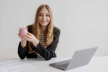 Gülümseyen genç bir kadın elinde pembe bir kumbara tutuyor. Yanında dizüstü bilgisayar ve evrak işleriyle bir masada oturuyor. Ev ofisinde tasarruf yapmanın önemini vurguluyor..