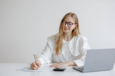 Gözlüklü genç bir kadın basit bir ofiste hesap makinesi kullanırken kağıda notlar yazar. Önünde açık duran bir dizüstü bilgisayar, bütçe işleriyle meşgul olduğunu gösteriyor..