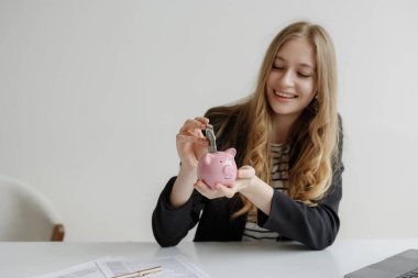 Genç bir kadın pembe domuz kumbarasına bozuk para yerleştirirken gülümsüyor. Finansal belgelerle dolu bir masada oturuyor. Biriktirmeye olan hevesini gösteriyor..