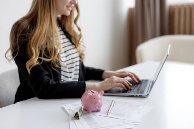 Beyaz bir masada oturan siyah ceketli bir kadın dizüstü bilgisayarda yazı yazıyor. Pembe domuz kumbarası ve finansal dökümanlar, bütçe planlama ve planlama faaliyetlerini gösteriyor..