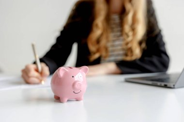 Bir kadın masanın başında oturmuş, pembe bir kumbara yakınlarda sergilenirken notlar yazıyor. Ayarlar, profesyonel bir ortamda mali planlama ve bütçe belirleme üzerinde durulmasını öneriyor.