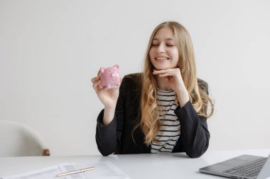 Uzun saçlı, siyah ceketli bir kadın parlak bir ofiste pembe bir kumbara gösteriyor. Evrak işleri ve dizüstü bilgisayarla çevrili finansal hedeflerini düşünürken memnun görünüyor..