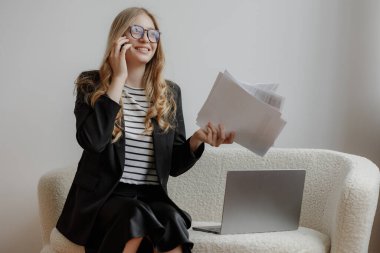 Akıllı kıyafetli genç bir kadın bir elinde belgelerle bir dizüstü bilgisayarın yanında otururken telefonuyla konuşuyor. Meşgul ve iyi aydınlatılmış bir çalışma alanına odaklanmış görünüyor..