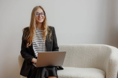 Genç bir kadın, dizüstü bilgisayarına odaklanmış modern bir kanepeye rahatça oturur. Gözlük takıyor ve çizgili bir gömlek giyiyor, rahat bir ofis ortamında profesyonel ama rahat bir hava yayıyor..