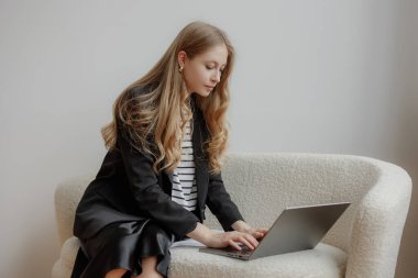 Uzun saçlı genç bir kadın, modern ofis ortamında şık beyaz bir koltuğa oturmuş, gündüz saatlerinde bir proje üzerinde özenle çalışıyor..