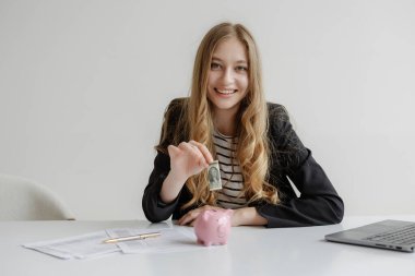 Uzun saçlı genç bir kadın çalışma yerindeki pembe domuz kumbarasına bir dolarlık banknot koyuyor. Finansal planlamayı gösteren belgeler ve dizüstü bilgisayarlar mevcut..