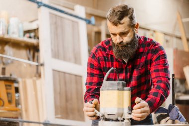 Becerikli bir marangoz ahşap bir parça üzerinde yönlendirici ile dikkatlice çalışır, alet ve malzemelerle dolu parlak bir atölyede işçilik sergiler..