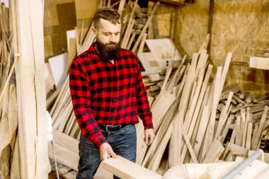 Ekose gömlek giymiş yetenekli bir zanaatkar hammadde dolu bir marangozhanedeki kereste parçasını inceliyor. Çevre kırsaldır, işçiliğin altını çizer..
