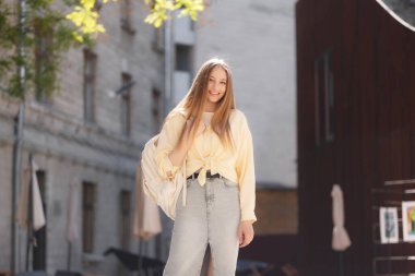 Genç bir kadın canlı bir şehir ortamında kendine güvenerek duruyor, gülümsüyor ve hafif günlük kıyafetler giyiyor. Güneş ışığı, dışarıda geçirdiği günün tadını çıkarırken rahat duruşunu aydınlatıyor..