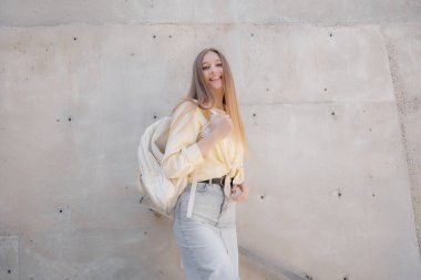 Genç bir kadın sırt çantası ve açık sarı bir bluz ve kot pantolon giyerek kendine güvenerek ayakta duruyor. Güneşli günün keyfini çıkarırken gülümsüyor, modern beton bir zemin tarafından çerçevelenmiş..