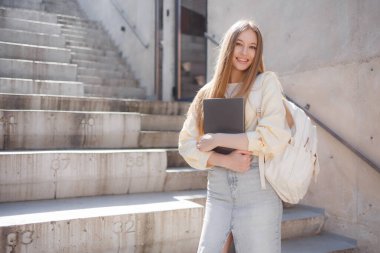 Genç bir kadın açık hava merdivenlerinde dizüstü bilgisayarını yakın tutarken gülümsüyor. Şık bir süveter ve kot etek giyiyor. Çalışma seansına hazır..