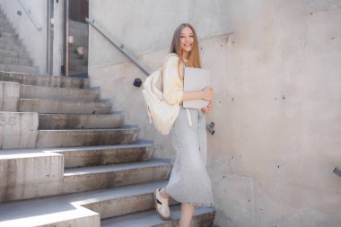 Çağdaş bir ortamda beton merdivenlerden çıkan genç bir kadın elinde bir dizüstü bilgisayar ve sıradan bir kıyafet ile kampüste günışığının tadını çıkarıyor..