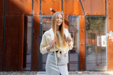 Genç bir kadın, sarı bluz ve kot etek giymiş çağdaş bir yapının önünde güvenle gülümsüyor, uzun saçları omuzlarından aşağı dökülüyor..