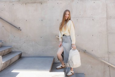 Uzun saçlı genç bir kadın şık beton merdivenleri tırmanıyor, hafif bir sırt çantası taşıyor. Rahat bir kıyafet giyiyor, bol bluzu ve kot eteği var, güneşli havanın tadını çıkarıyor..