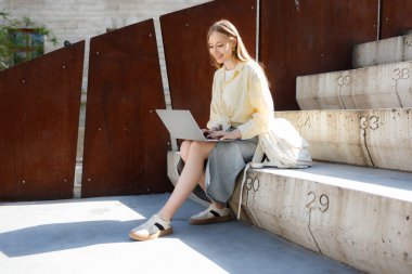 Genç bir kadın dışarıda merdivenlerde otururken dizüstü bilgisayarında yazıyor. Açık sarı bir bluz ve gri bir etek giyiyor. Şehir ortamında güneşli bir günün tadını çıkarıyor..