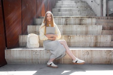 Çağdaş bir ortamda beton merdivenlerde oturan uzun saçlı genç bir kadın elinde dizüstü bilgisayar ve sırt çantasıyla derslerine hazırlanırken gülümsüyor. Güneş ışığı parlıyor..