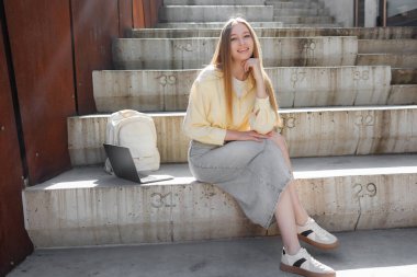 Genç bir kadın, sarı bluz ve kot etek giymiş, beton merdivenlerde oturuyor. Yanında bir laptop var ve güneşli havanın tadını çıkarıyor, neşeli ve rahat görünüyor..