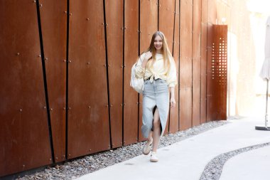 Uzun saçlı bir kadın, sıradan ama modaya uygun kıyafetlerini sergileyerek uzun bir yolda yürüyor. Modern bina arkaplanı, o anın kentsel atmosferini arttırır..