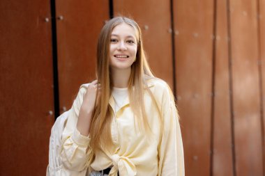 Uzun sarı saçlı genç bir kadın kahverengi bir duvarın önünde dururken gülümsüyor. Açık sarı bir gömlek giyiyor ve neşeli bir hava saçan bir sırt çantası taşıyor..