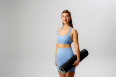 Parlak, geniş bir stüdyo ortamında şık spor giysileri içinde yoga minderi tutan genç bir kadın canlandırıcı bir yoga seansına hazırlanıyor..