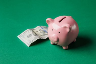 Yeşil bir yüzey üzerinde katlanmış bir banknotun yanında duran pembe bir kumbara tasarruf ve mali planlama kavramını vurguluyor. Neşeli tasarım dikkatleri kişisel finansmana çekiyor..