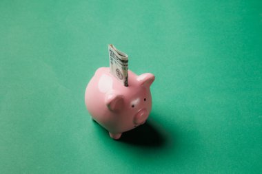 Pembe bir kumbara, yuvasından sarkan bir dolarlık banknotla canlı yeşil bir zemin üzerinde oturur. Bu görsel tasarruf ve finansal planlamayı vurguluyor.