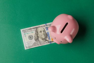 Parlak yeşil bir yüzey üzerinde pembe bir domuz kumbarası 100 dolarlık banknotun yanında duruyor. Elementler tasarruf ve mali planlamayı simgeliyor.