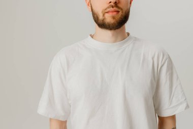 Genç bir adam, minimalist bir stili vurgulayan basit beyaz bir tişört giyerek kendinden emin bir şekilde ayakta duruyor. Tarafsız zemin, sakin bir atmosfer yaratarak gündelik modanın altını çiziyor..