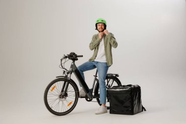 Bir adam elektrikli bisikletle teslimat vardiyasına hazırlanıyor. Yeşil bir kask takıyor ve yanında minimalist bir şehir stüdyosunda yalıtımlı bir teslimat çantası var..