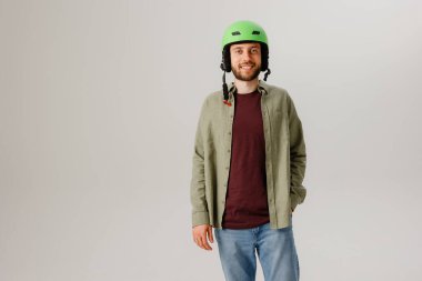 Parlak yeşil bir kask takan genç bir adam, kestane rengi bir gömlek ve açık renk bir ceketten oluşan günlük bir kıyafet giyerek gülümsüyor. Sade bir geçmişi olan minimalist bir stüdyoda duruyor..