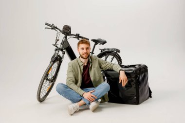 Genç bir adam parlak bir stüdyo ortamında bisiklet ve teslimat çantasının yanında tesadüfen yerde oturuyor, kuryelik işi için hazırlanıyor. Rahatlamış ve harekete hazır görünüyor..