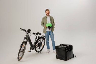 Bir teslimatçı elinde yeşil bir miğfer olan elektrikli bir bisikletin yanında duruyor. Siyah bir teslimat çantası yerde duruyor, binicilerin ilerideki işe hazır olduklarını vurguluyor..