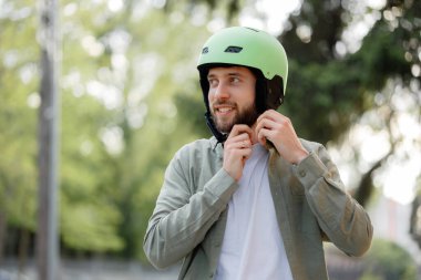 Genç bir adam yeşil bisiklet kaskını ayarlıyor, mutlu bir şekilde parkta gülümsüyor. Ağaçlar, sıcak bir öğleden sonra bisiklet macerasına hazırlanırken canlı bir arka plan sağlar..