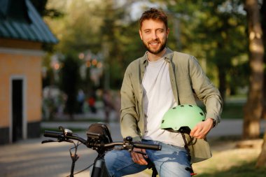 Neşeli bir adam elinde yeşil bir miğferle bisikletinin üzerinde oturur, öğleden sonra yeşil ve sıcak güneş ışığıyla çevrili, canlı bir parkta gezintiye çıkmaya hazırdır..