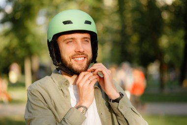 Genç bir adam canlı bir parkta yeşil kask takıyor. Ağaçlarla çevrili neşeli bir ifadesi var ve açık havada havanın tadını çıkaran insanlar..